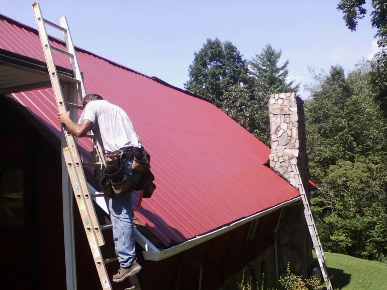 Roofer on a ladder beside a red metal roof and stone chimney — Soffit & Fascia Repair by Roofing Brothers in Brockway